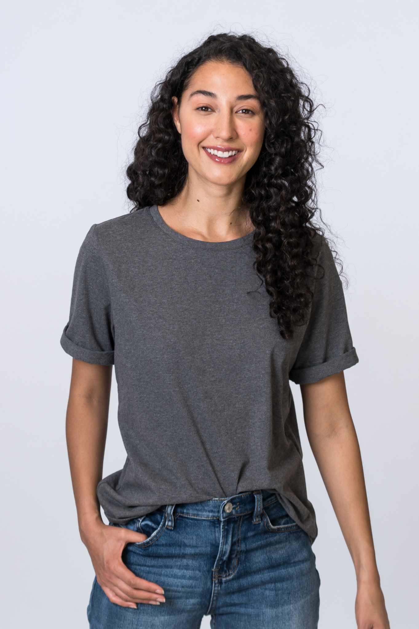 Woman smiling, wearing a dark gray, cuffed sleeve short sleeve top tucked into medium wash jeans. Front of clothing is being shown