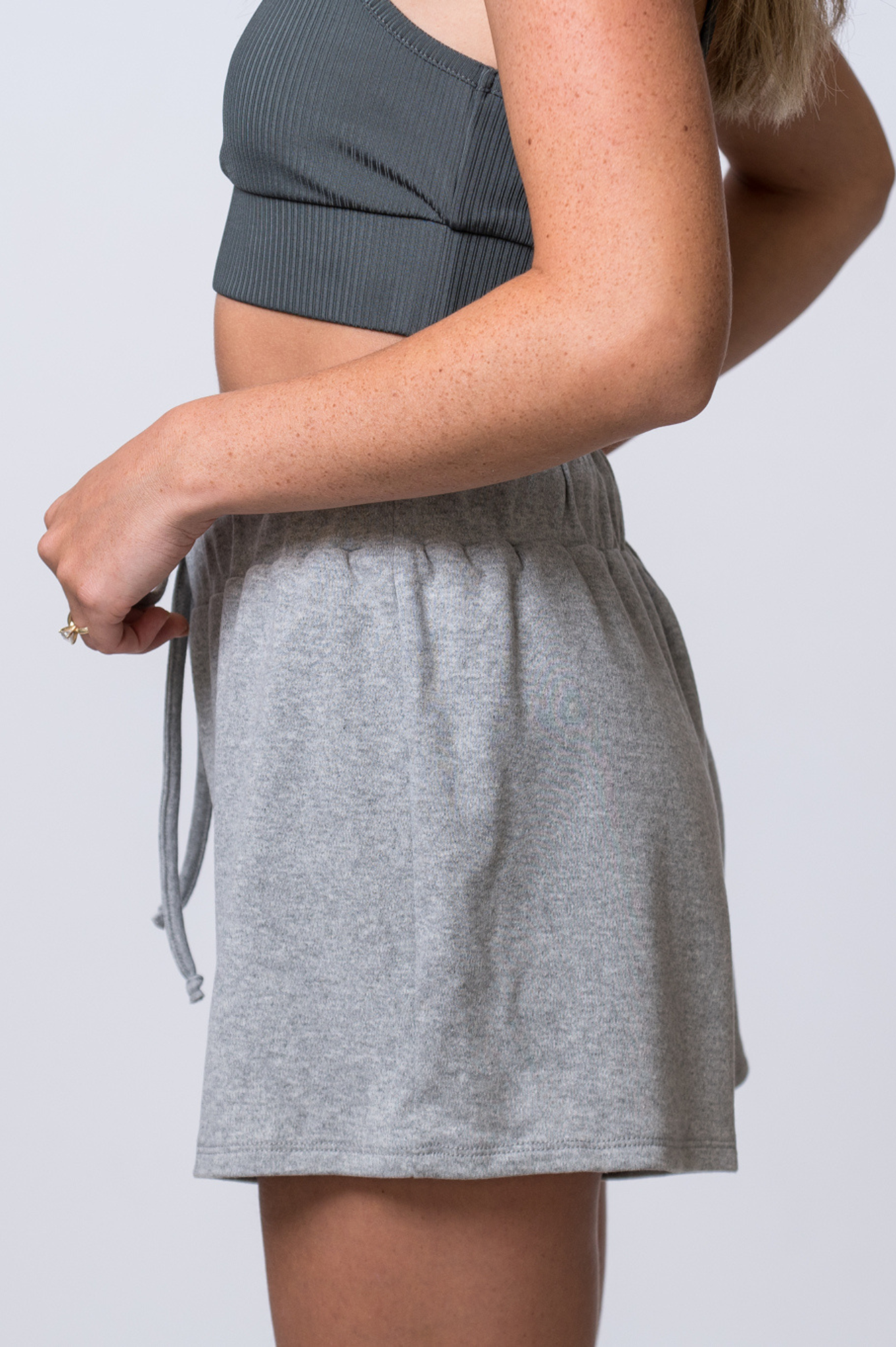 Woman wearing light gray, drawstring sweat shorts and a dark gray, ribbed sports bra. Side of clothing is being shown