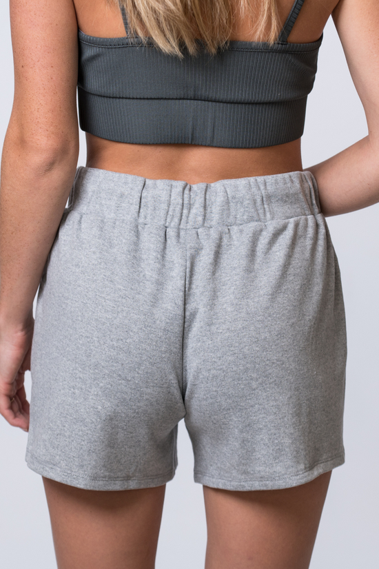 Woman wearing light gray, drawstring sweat shorts and a dark gray, ribbed sports bra. Back of clothing is being shown