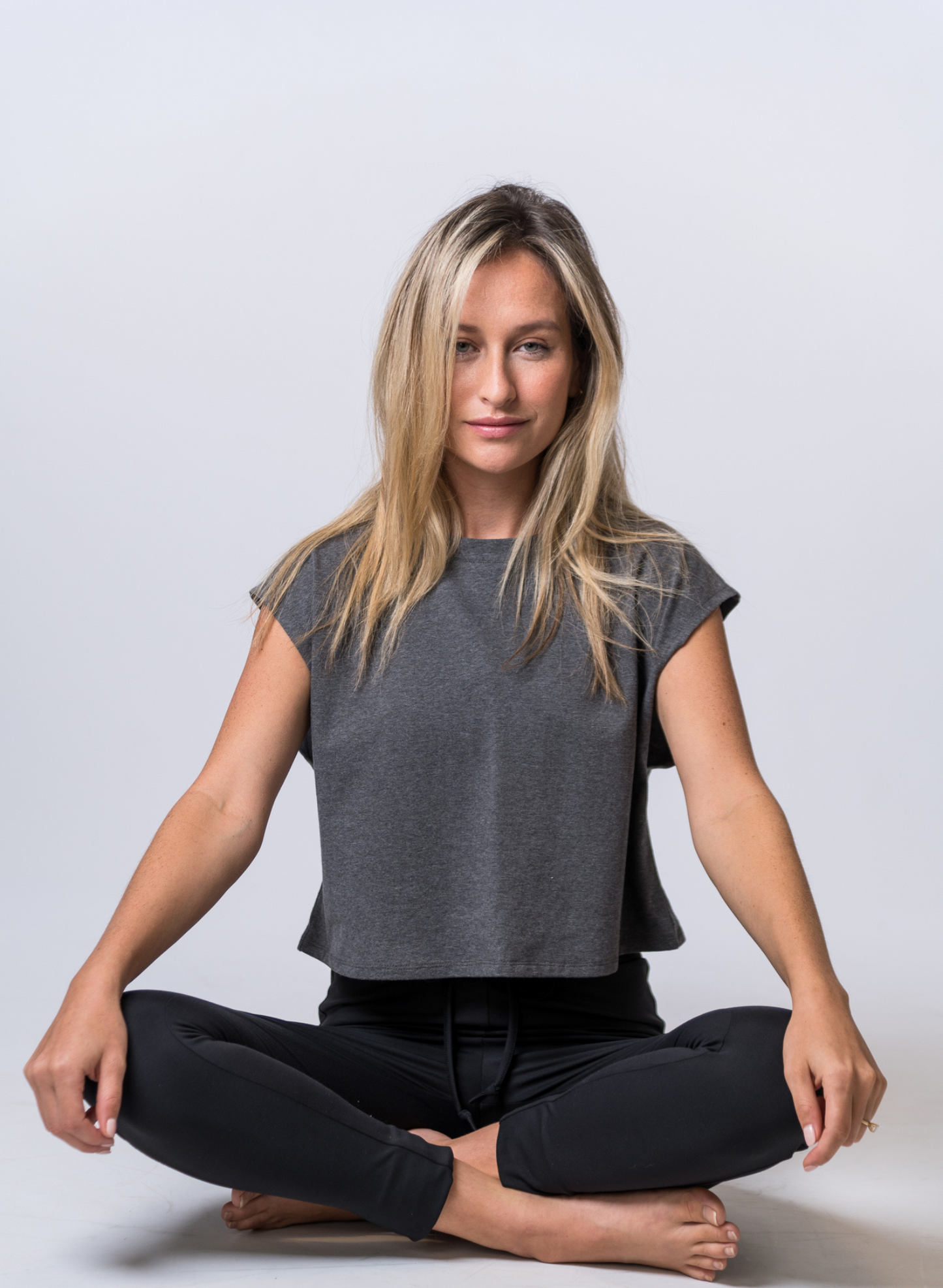 Woman sitting with her legs crossed, wearing a dark gray, crop top with black drawstring lounge pants. Front of clothing is being shown