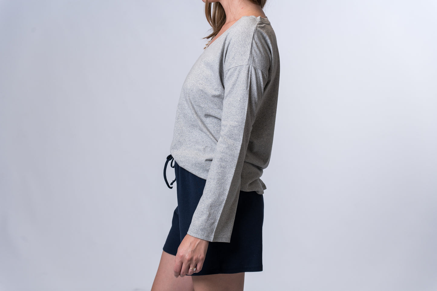 Woman wearing a gray, long sleeve lounge top with navy blue drawstring lounge shorts. Side of clothing is being shown