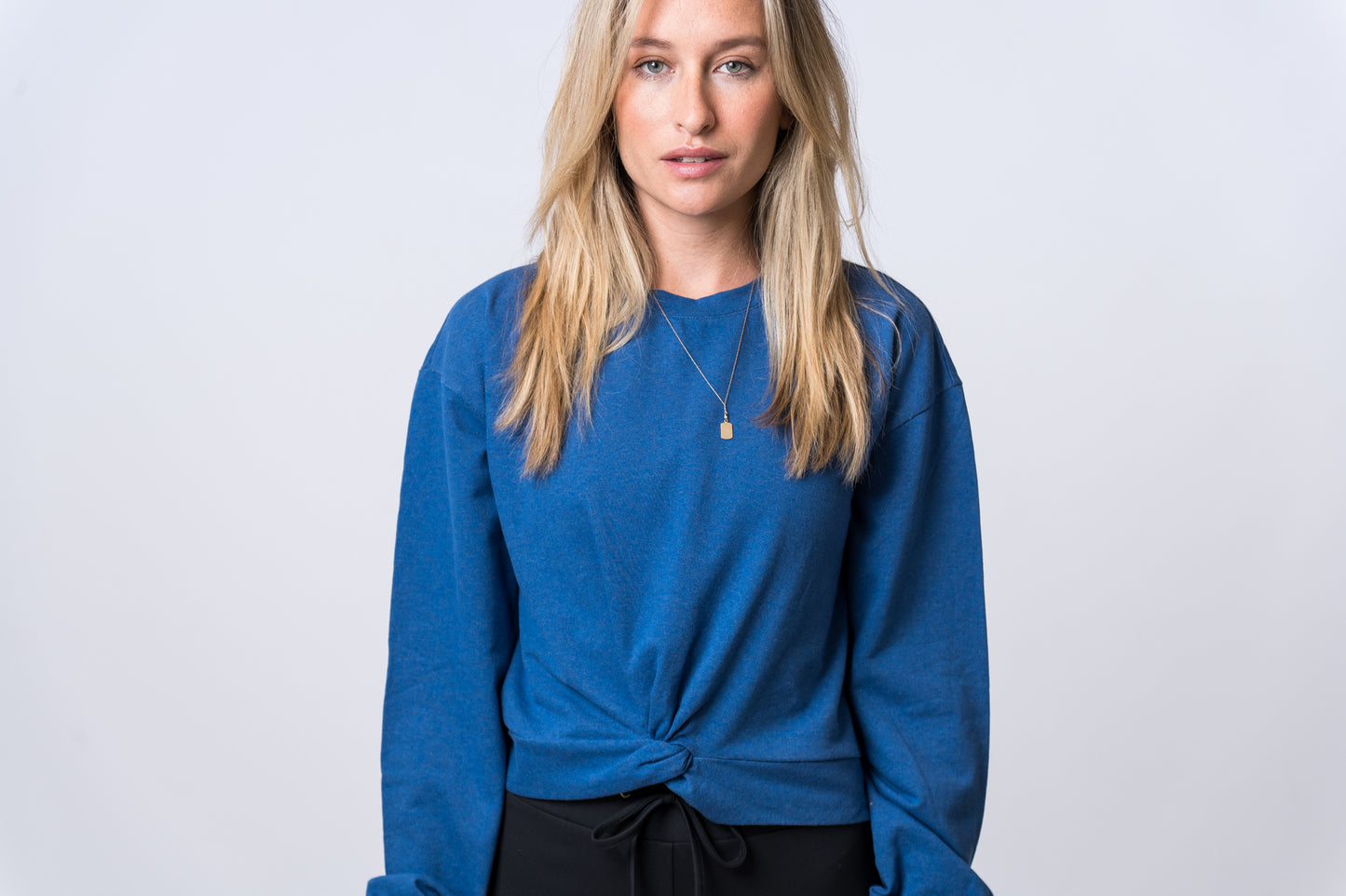 Woman wearing a blue sweatshirt with knot and black drawstring lounge pants. Front of clothing is being shown