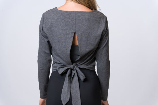 Woman wearing a dark gray, long sleeve top with open back and tie with black drawstring pants. Back of clothing is being shown