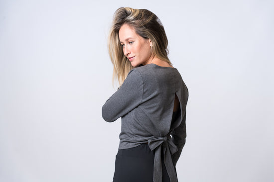 Woman wearing a dark gray, long sleeve top with open back and tie with black drawstring pants. Side and back of clothing is being shown