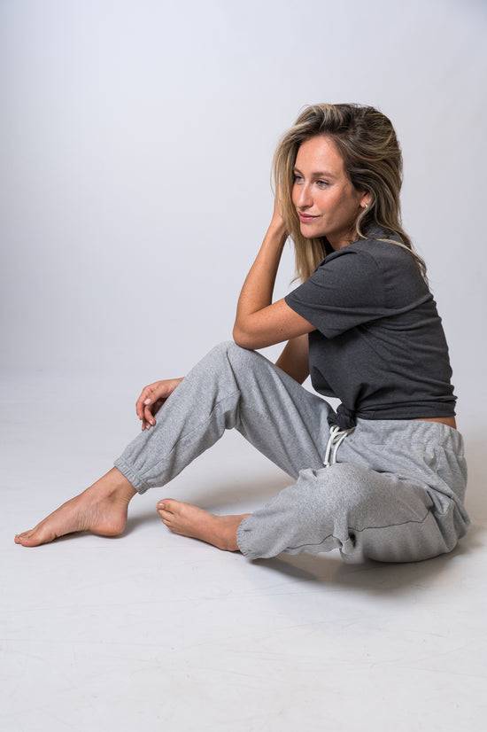 Load image into Gallery viewer, Woman sitting, wearing light gray, drawstring sweatpants rolled at the bottom, with a dark gray, tie knot top. Front and side of clothing is being shown

