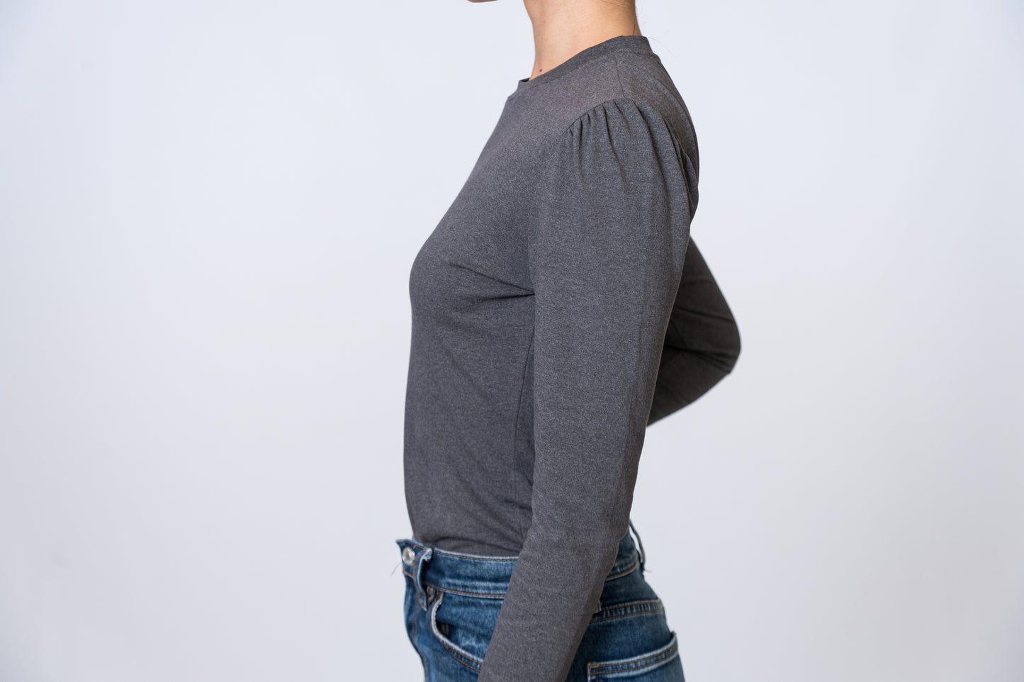 Woman wearing a gray, long sleeve top with puff sleeves and medium wash jeans. Side of clothing is being shown