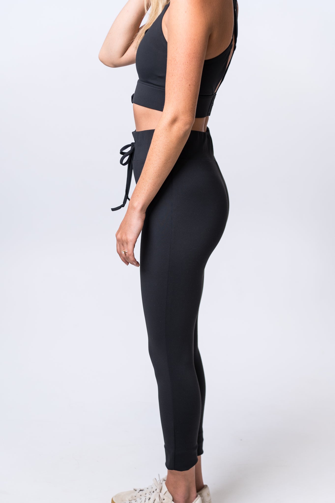 Woman wearing black, ankle length drawstring leggings with matching sports bra and white sneakers. Side of clothing is being shown